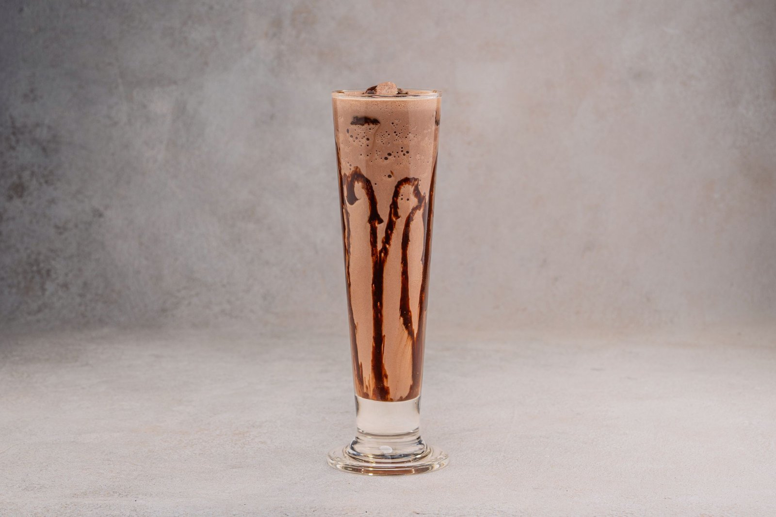 Creamy chocolate milkshake in a tall glass with chocolate drizzle on a neutral background.